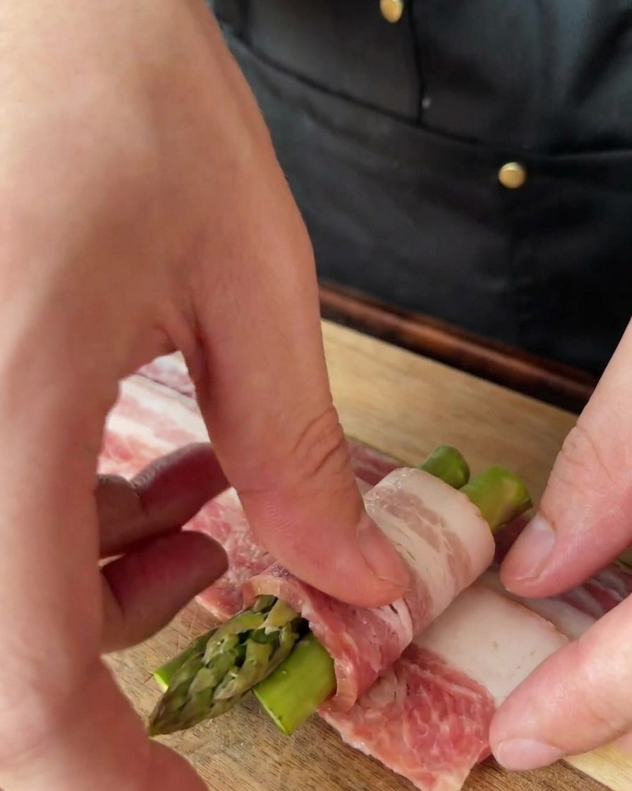 Mains enroulant une fine tranche de bacon autour dun fagot de trois asperges sur une planche en bois.