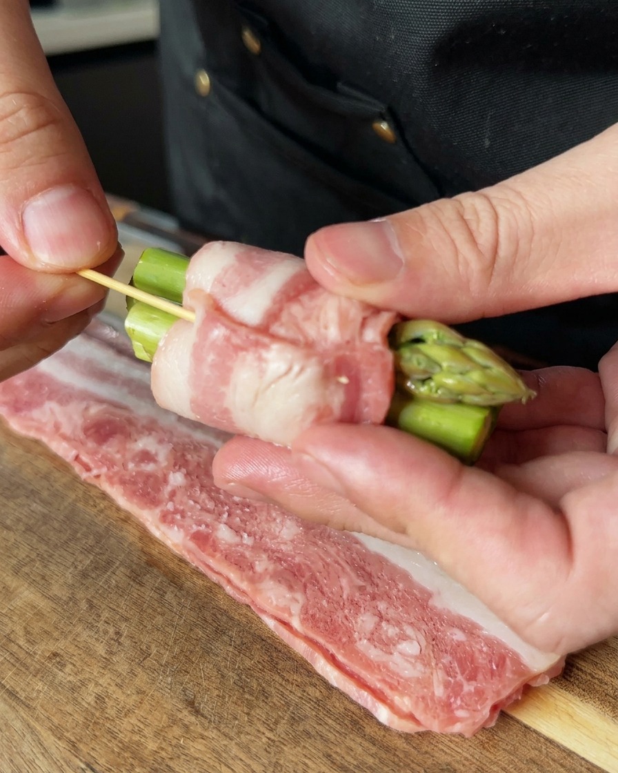 Une main utilisant un cure-dent en bois pour fixer un fagot dasperges au bacon.