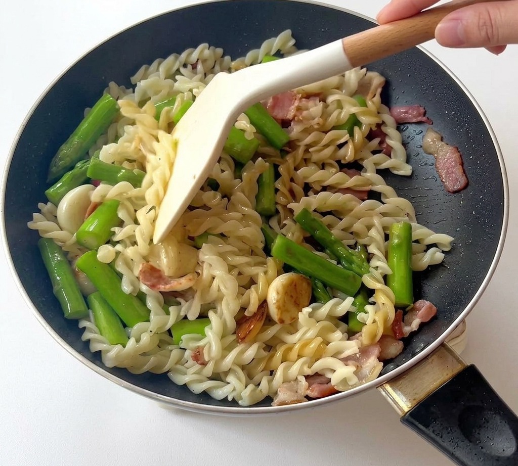 Spatule mélangeant des fusilli, des asperges et du bacon dans une poêle noire.