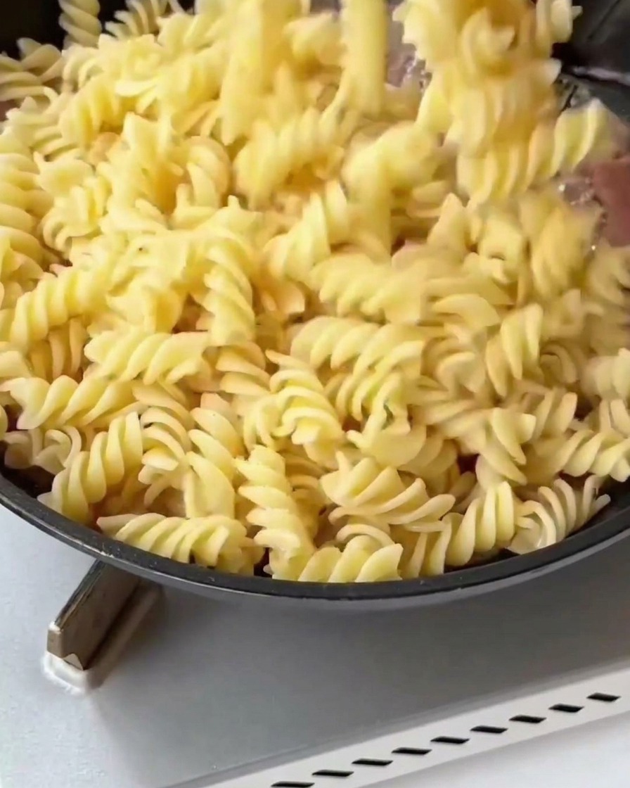 Fusilli cuits et asperges blanchies ajoutés dans une poêle avec du bacon et des oignons dorés.