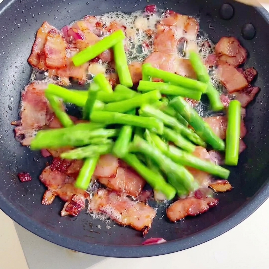 Une poêle contenant des morceaux de bacon croustillants et des asperges vertes fraîches, rejoints par des fusillis cuits.