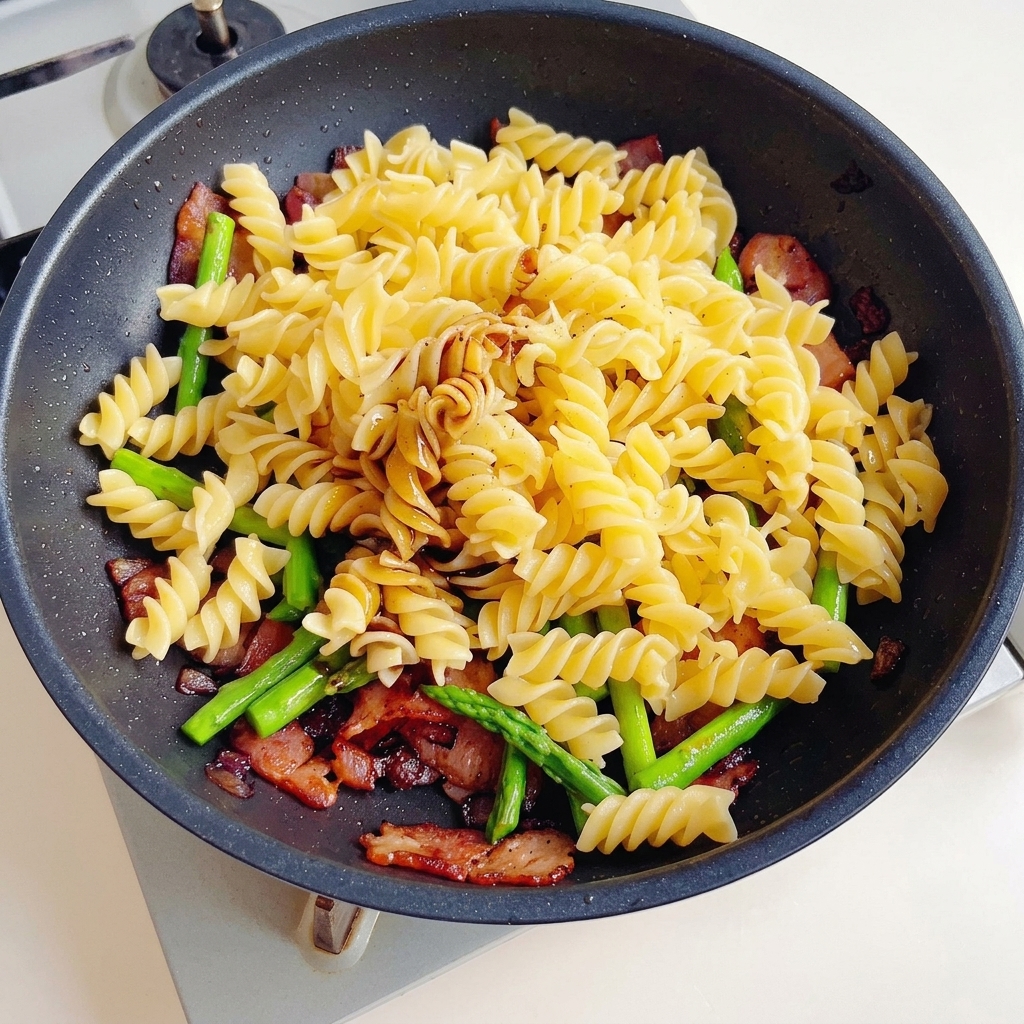 Sauce soja, sauce soja noire et sauce aux huîtres versées sur un mélange de pâtes, bacon et asperges dans une poêle antiadhésive.
