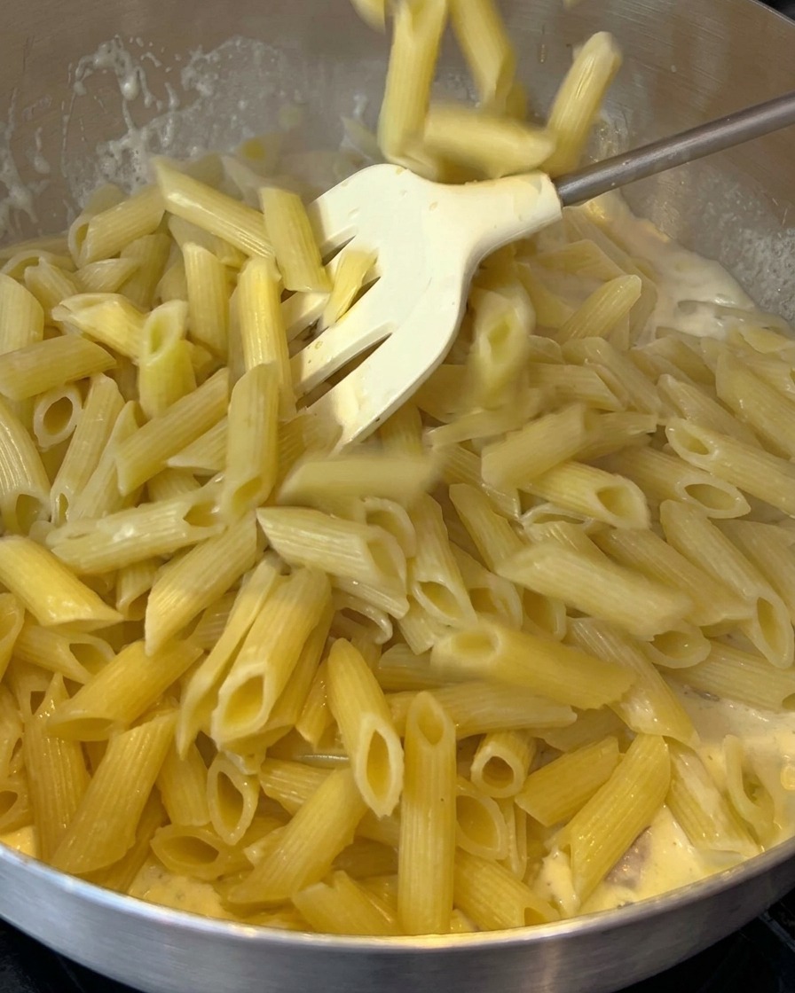Des penne cuites ajoutées dans une poêle avec une sauce crémeuse au bacon.