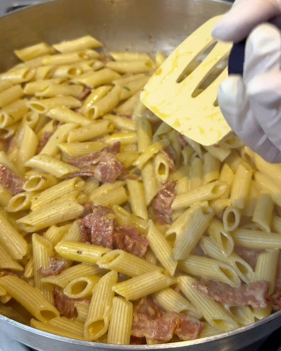 Du sel au citron moulu au-dessus dune poêle de penne crémeuses en train dêtre mélangées.