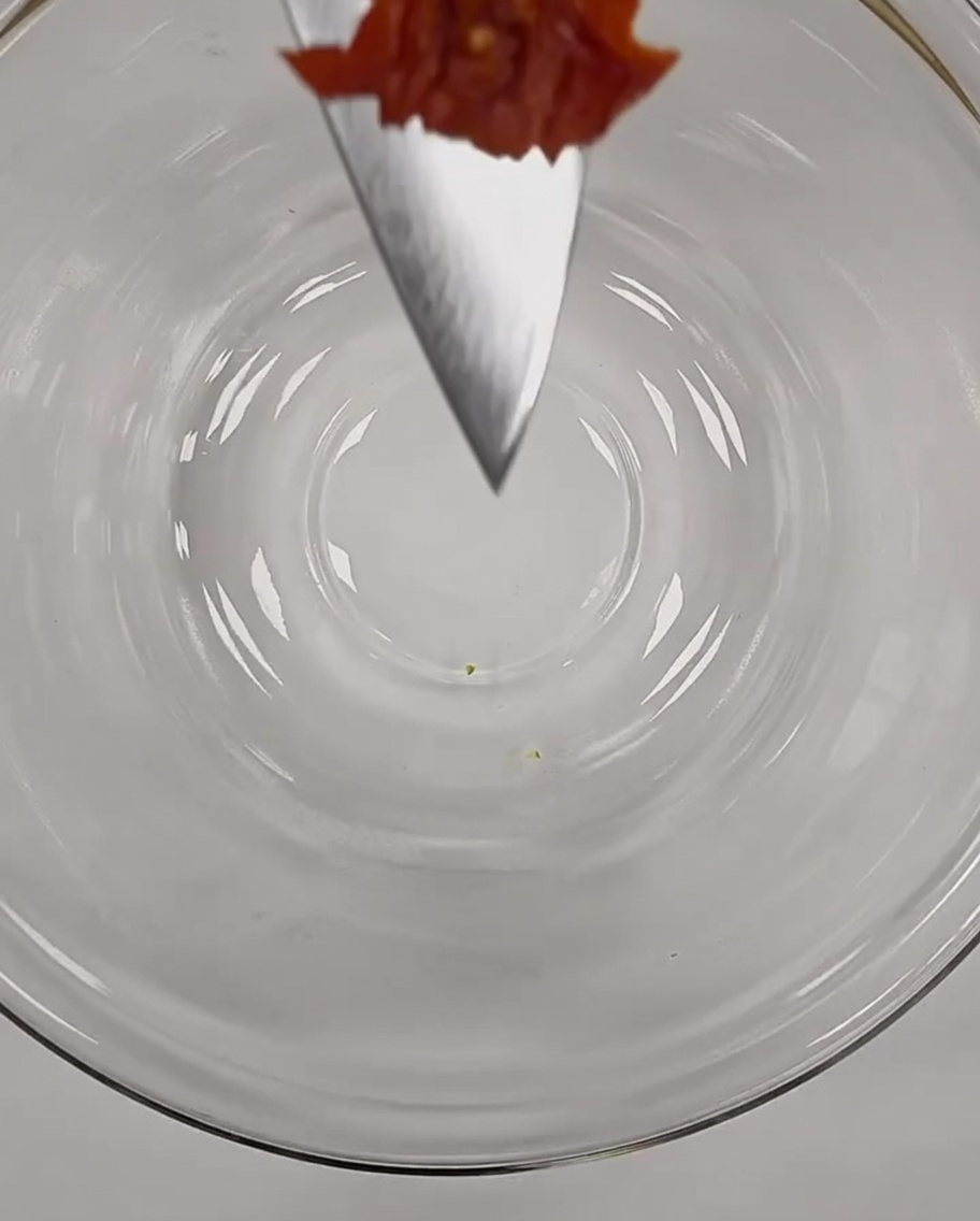 Un petit morceau de tomate séchée finement haché, délicatement équilibré sur la pointe dun couteau de chef au-dessus dun bol en verre.