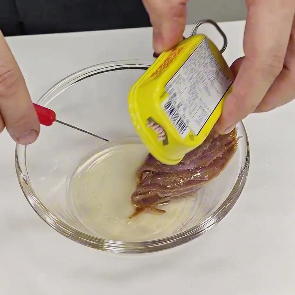 Des mains retirant soigneusement un filet danchois dune boîte en métal jaune pour le placer dans un petit bol de préparation en verre transparent.