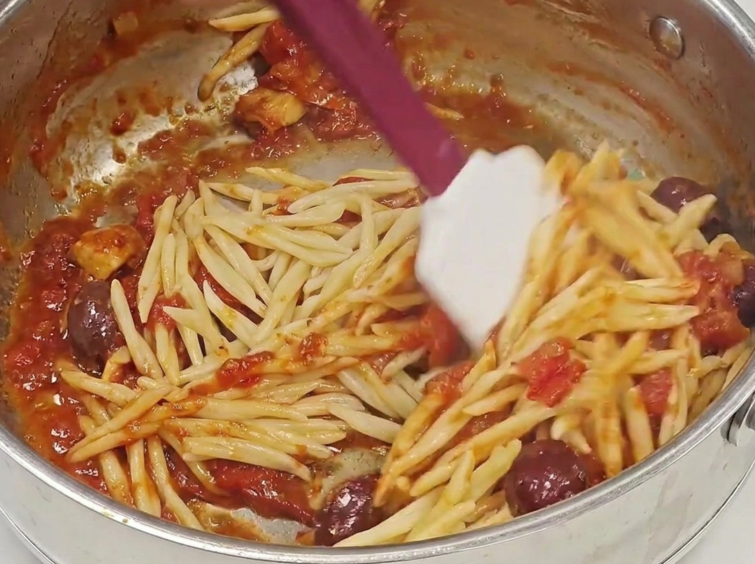 Des pâtes Trofie égouttées ajoutées dans une poêle remplie dune riche sauce Puttanesca rouge.