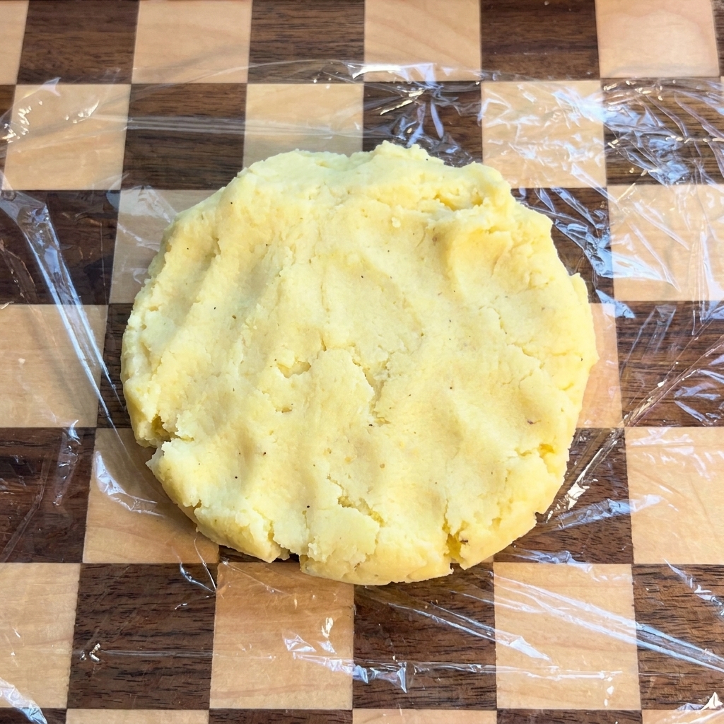 Une galette de pomme de terre plate et circulaire reposant sur une planche à découper à damier recouverte de film plastique.