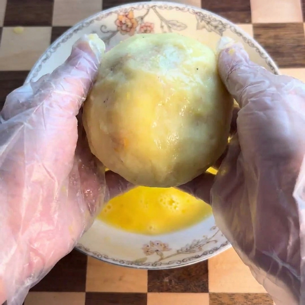 Une boule de pomme de terre trempée et roulée dans une assiette creuse contenant de lœuf jaune battu.