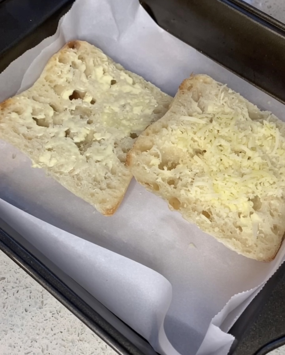 Du parmesan râpé saupoudré sur les tranches de ciabatta beurrées sur une plaque.