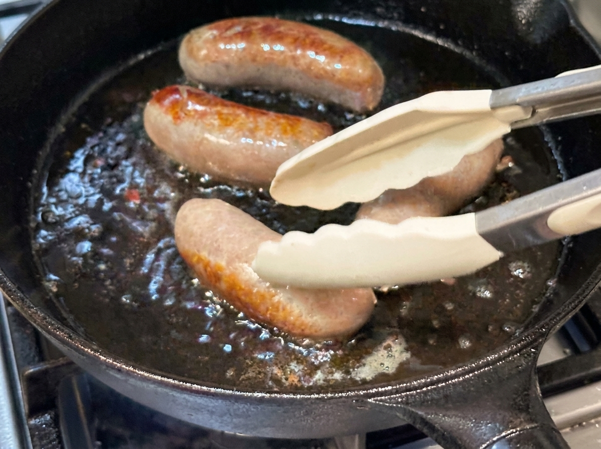 Saucisses entières retournées avec des pinces en silicone dans une poêle en fonte chaude.