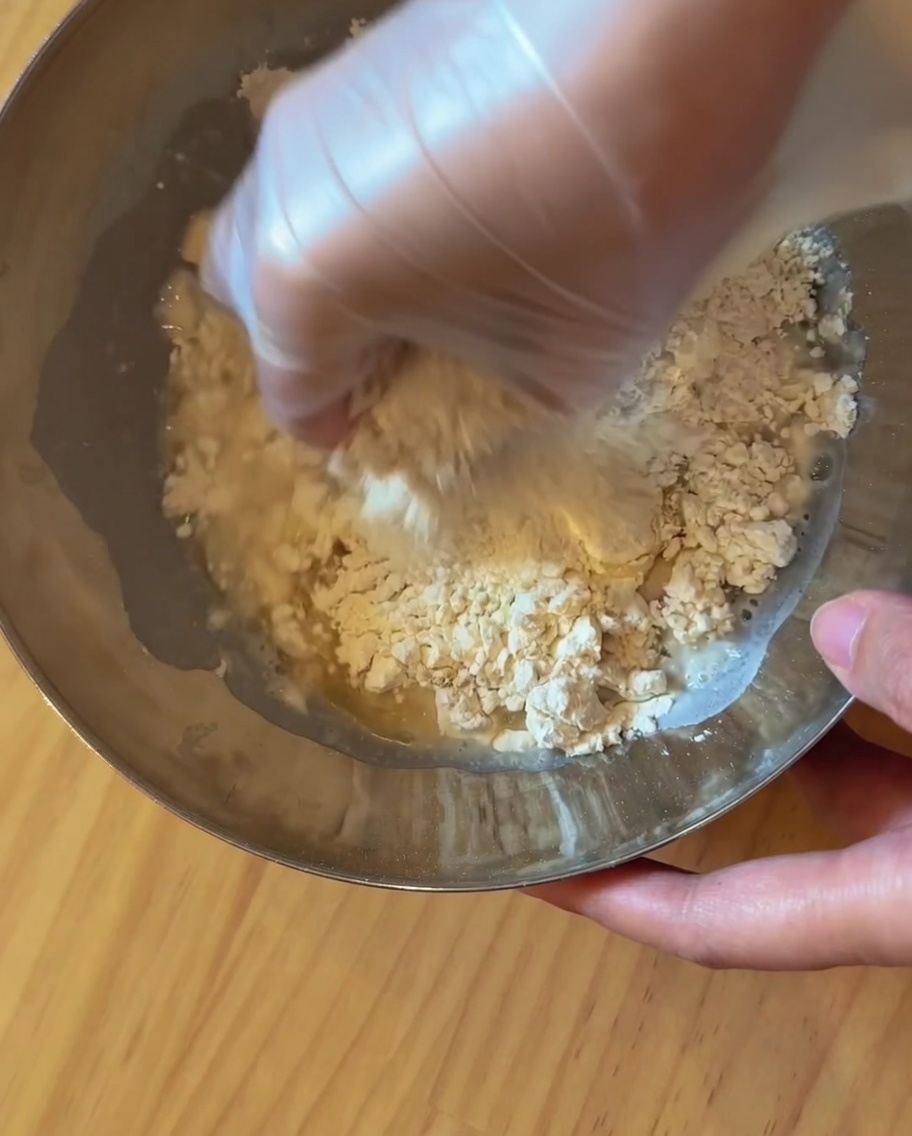 Une main portant un gant en plastique pétrissant une boule de pâte dans un bol en métal.