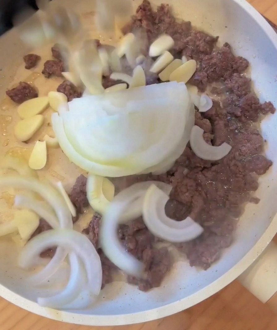 Des oignons émincés et de lail ajoutés dans une poêle avec la viande hachée dorée.