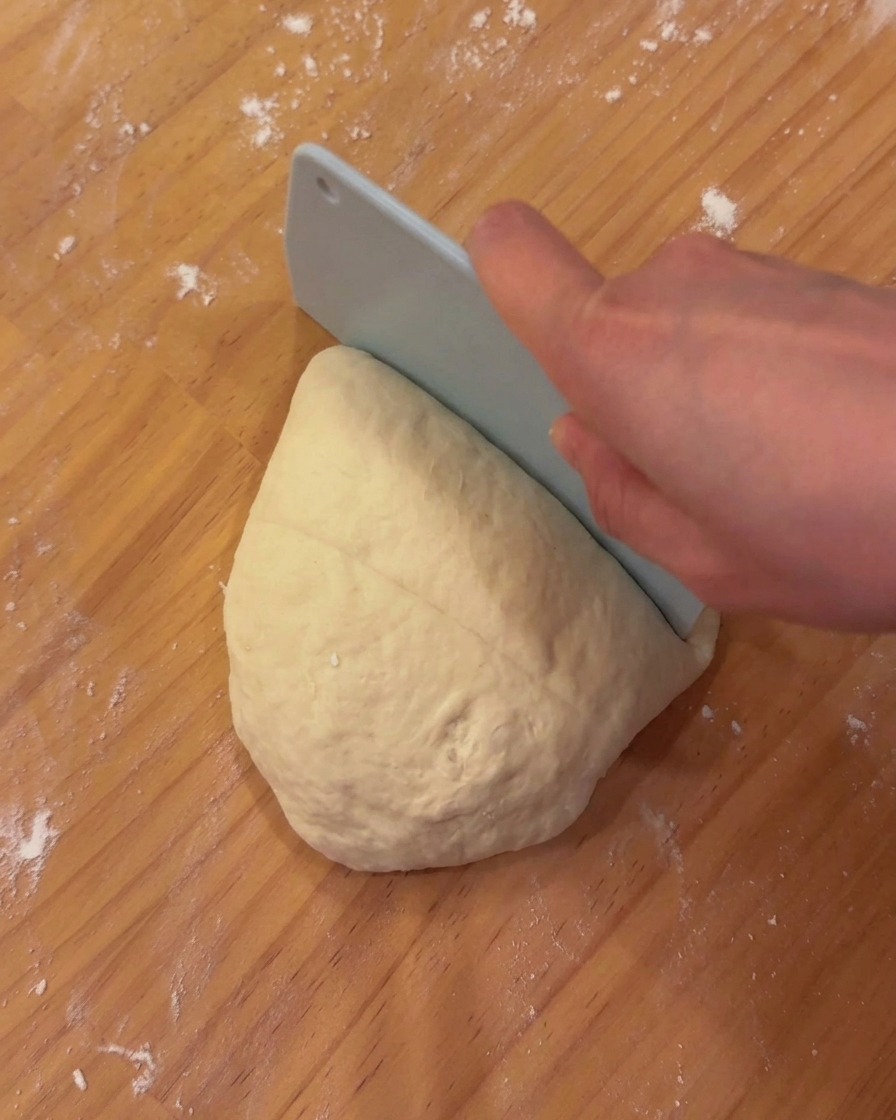 Une masse de pâte à pizza fermentée et lisse reposant sur une planche en bois avec un coupe-pâte.
