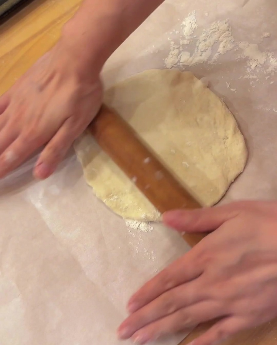 Un rouleau à pâtisserie en bois aplatissant un morceau de pâte à pizza rond sur du papier sulfurisé.