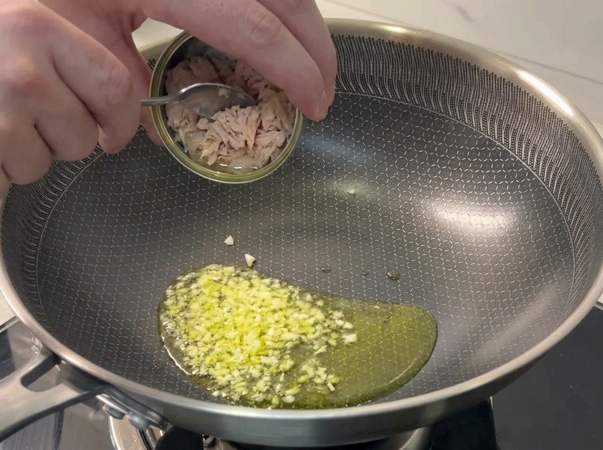 Ajout de morceaux de thon à la cuillère depuis une petite boîte dans une poêle avec de lail grésillant.