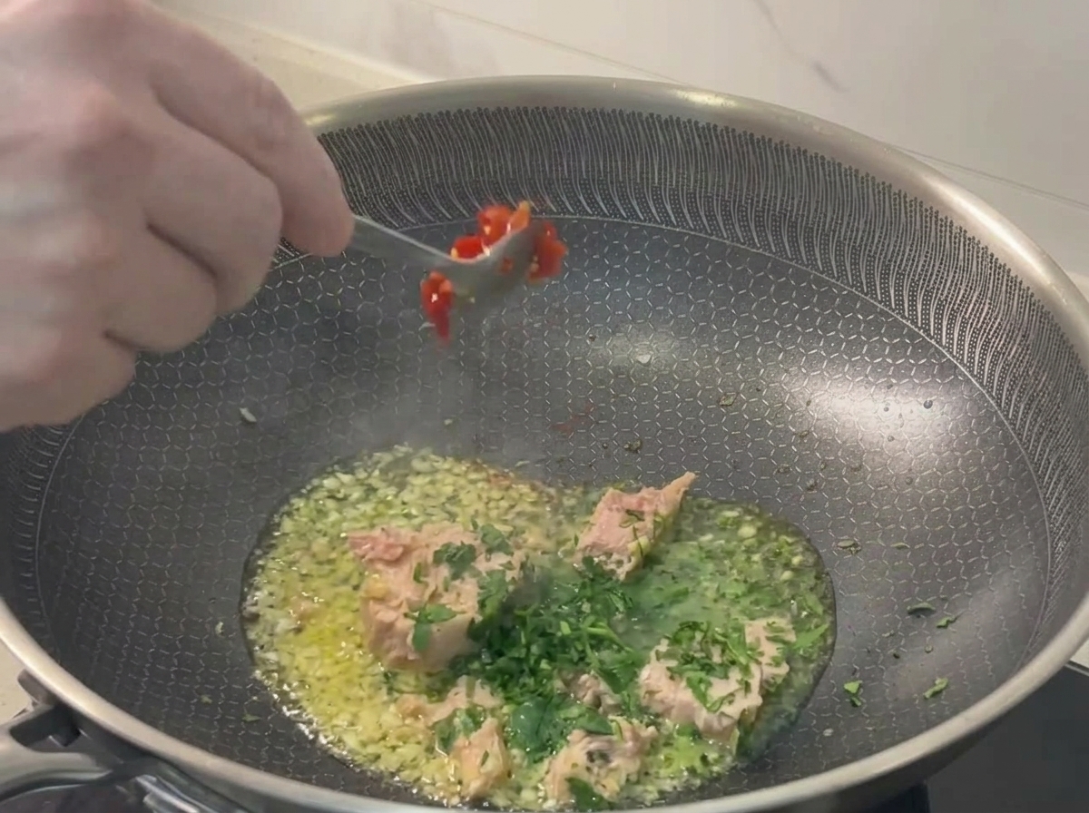 Une cuillère en métal ajoutant du piment rouge haché dans la poêle contenant le thon, lail et le persil.