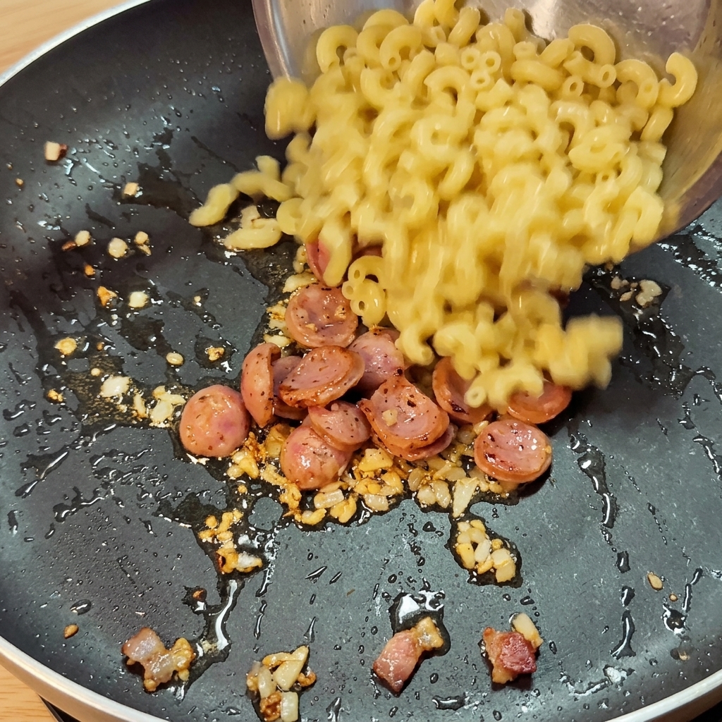 Des macaronis cuits versés dans une poêle contenant les tranches de saucisse sautées, les aromates et la sauce épicée.