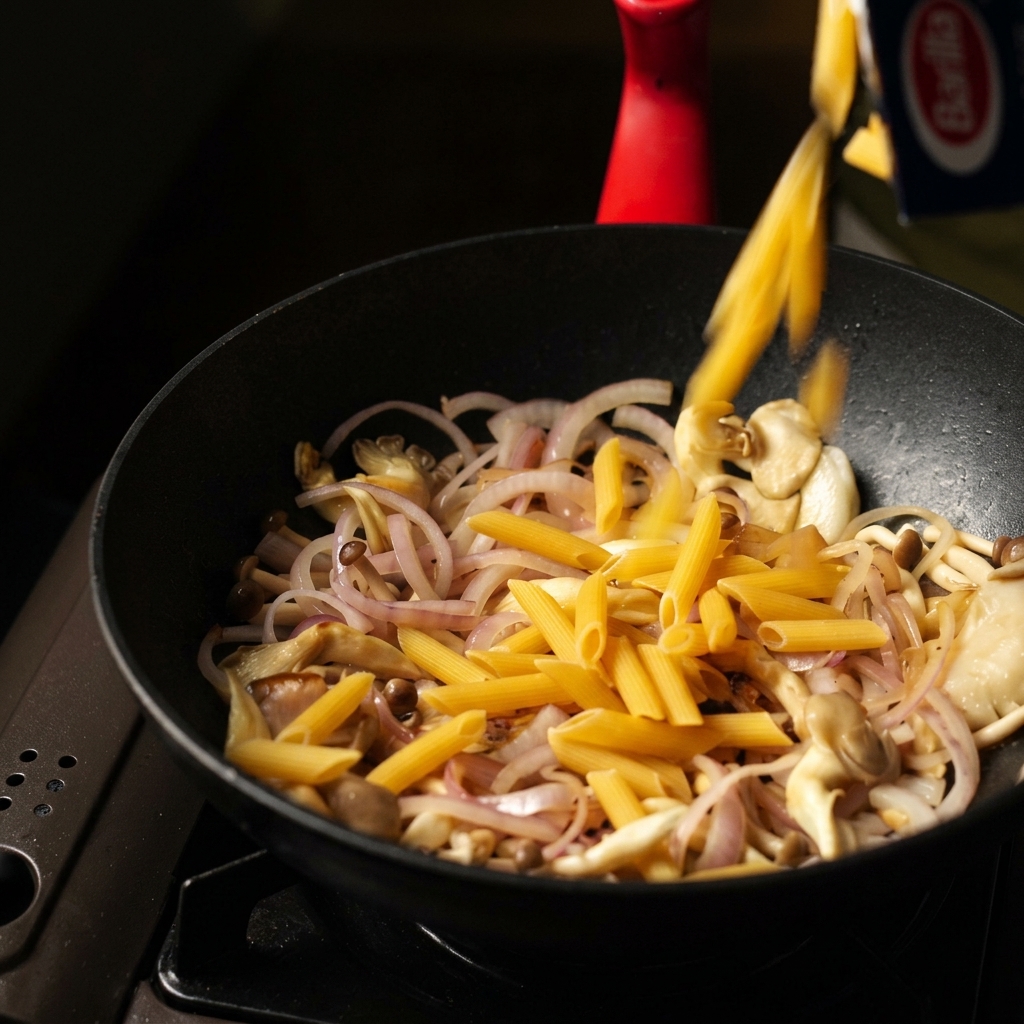 Des pennes crues versées depuis une boîte dans une sauteuse noire contenant les champignons et oignons revenus.