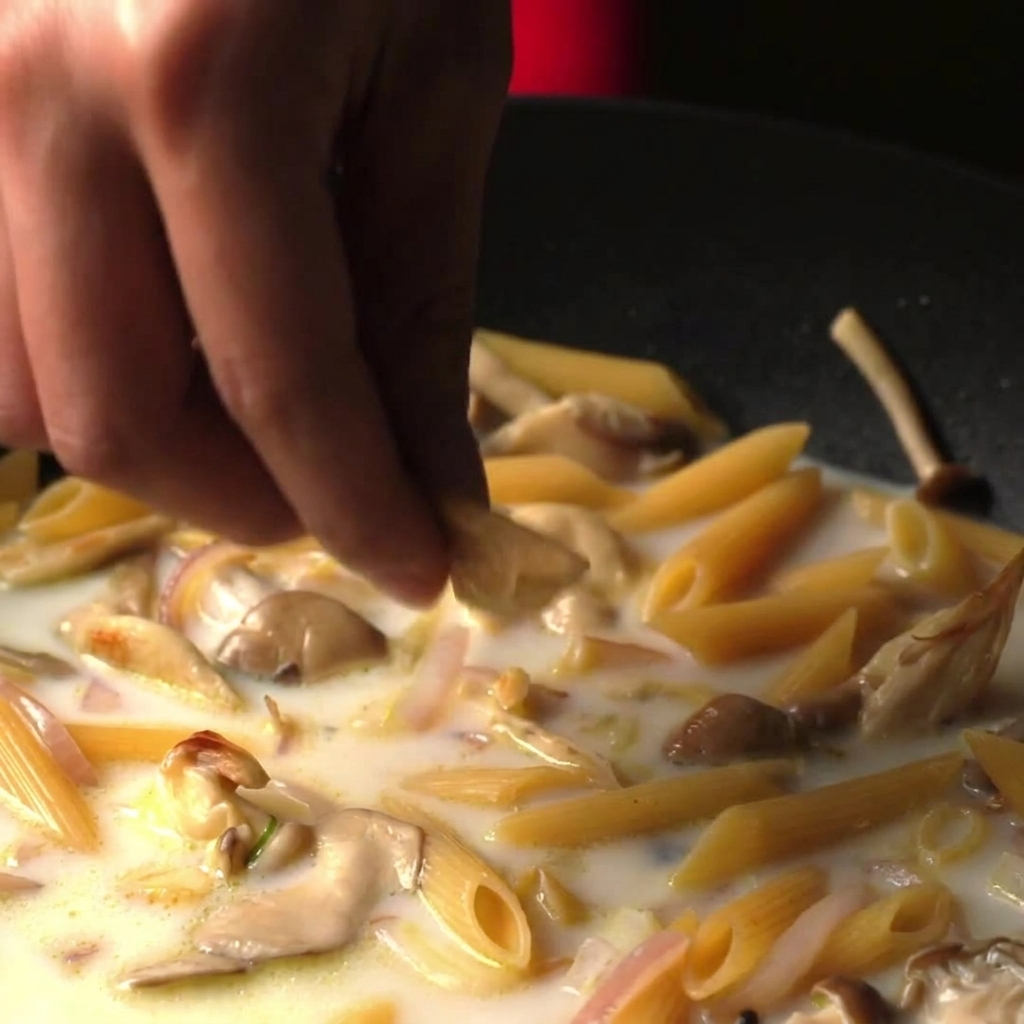 Des doigts émiettant un bouillon cube dans une sauteuse remplie de pennes mijotant dans un bouillon blanc et crémeux.