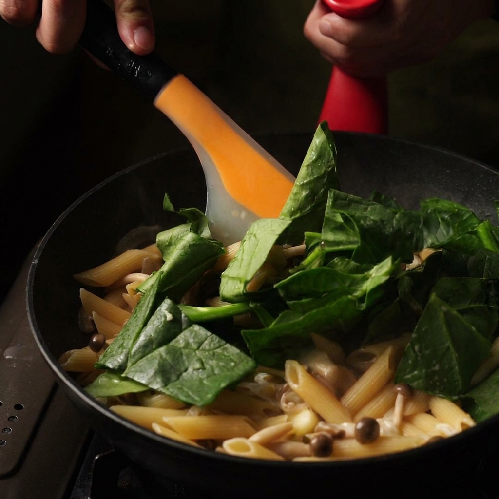 Une spatule orange mélangeant des feuilles dépinards bien vertes et du beurre dans une sauteuse de pennes crémeuses.