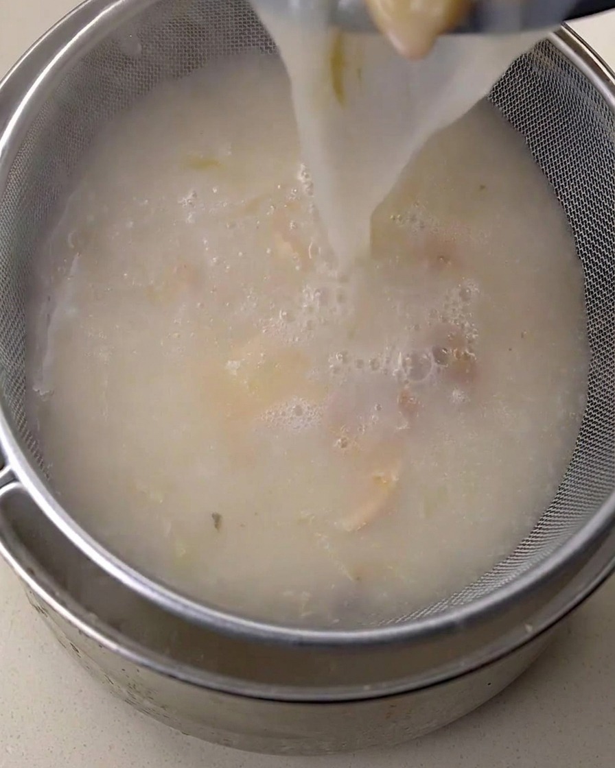 Bouillon de poulet blanc crémeux versé à travers un tamis fin dans un récipient métallique.