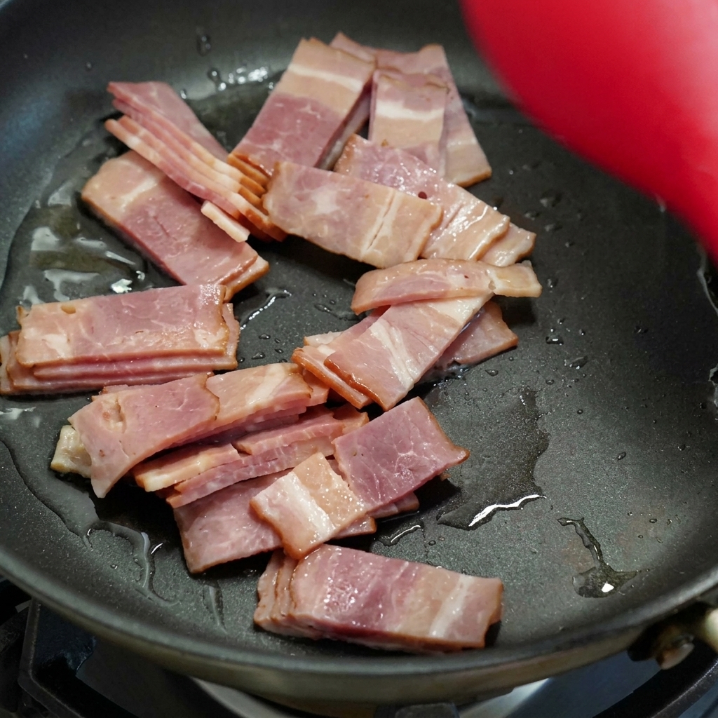 Tranches de bacon en train de cuire et de dorer dans une poêle noire, remuées avec une spatule rouge.