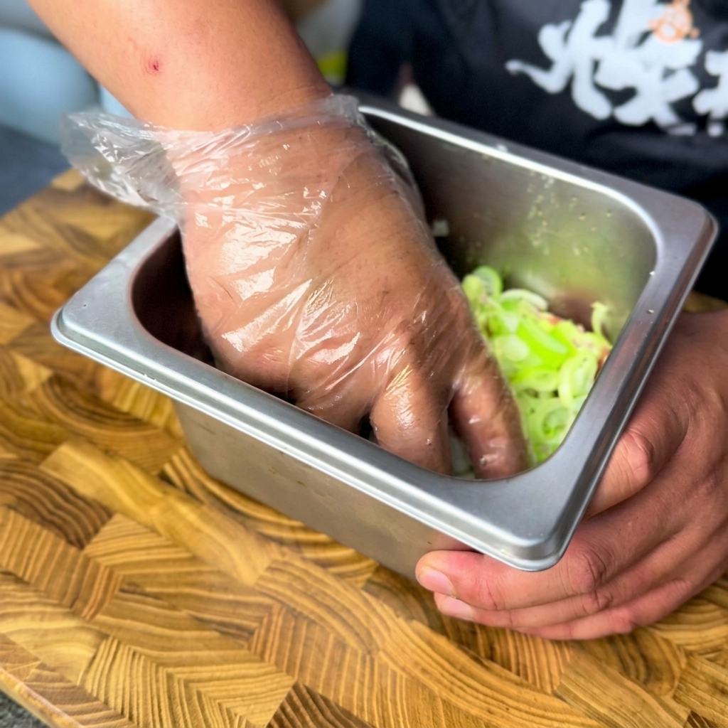 Une main gantée mélangeant des oignons verts hachés et du bœuf mariné dans un récipient en métal.