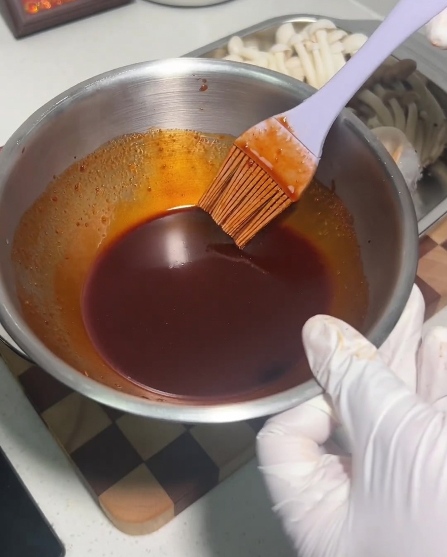 Un bol en métal contenant un mélange de sauce sombre et épicée remué avec un pinceau en silicone.