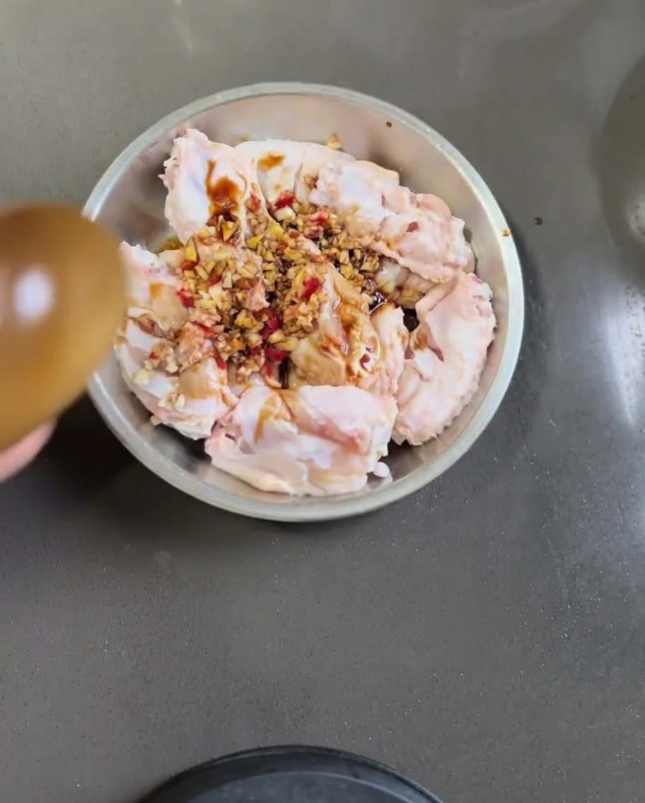 Ailes de poulet crues dans un bol en métal mélangées avec de lail, du piment, de la sauce soja et de lhuile de sésame.