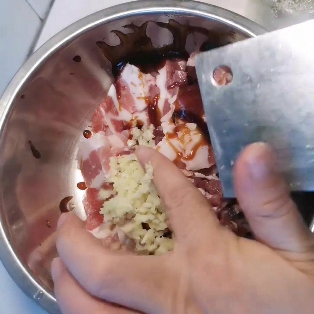 Un bol en métal contenant de la poitrine de porc en tranches, assaisonnée dail haché et de diverses sauces.