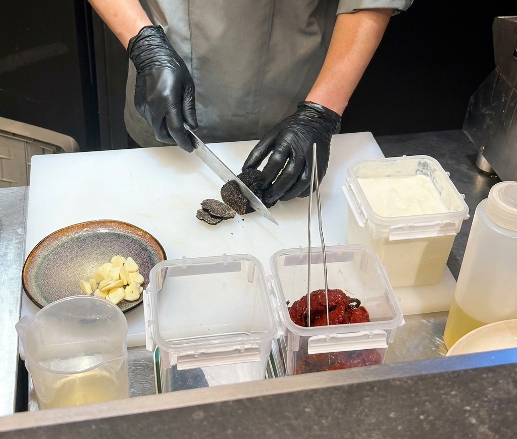 Un chef portant des gants noirs hache des piments noirs séchés sur une planche à découper blanche.