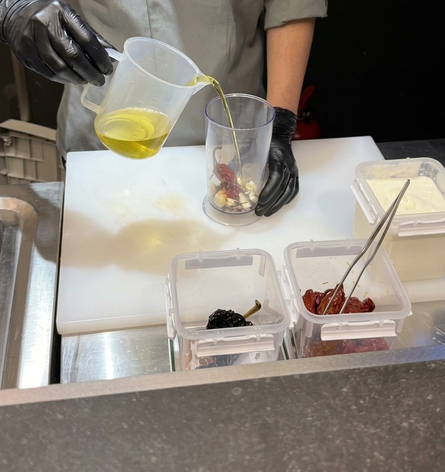 Un chef verse un liquide clair dans un grand bol mixeur contenant de lail et des piments.