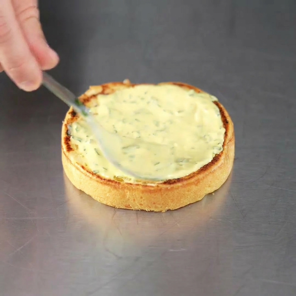 Un pain à burger grillé nappé de sauce verte aux herbes, prêt à recevoir le steak.