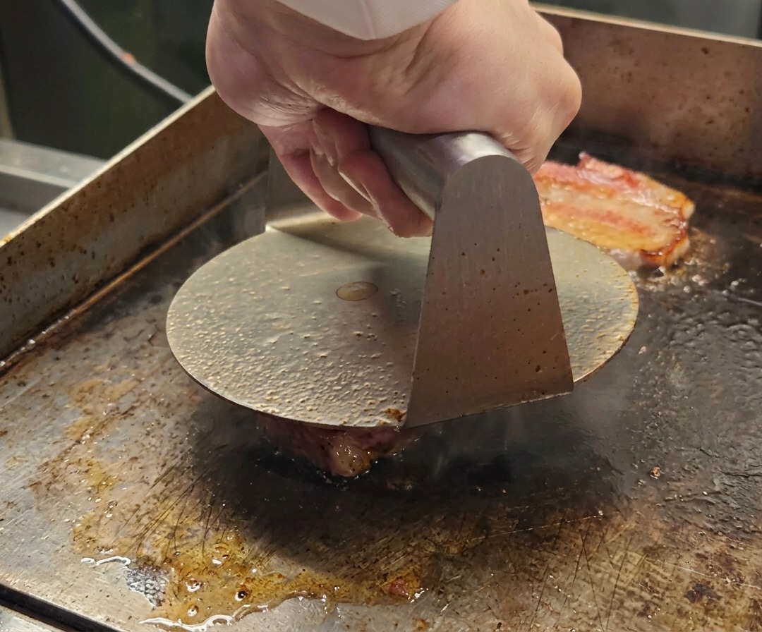 Une presse à burger métallique aplatissant une boule de bœuf Wagyu haché sur une surface de cuisson chaude.