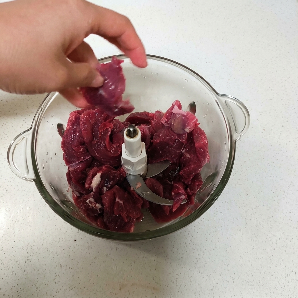 Une main plaçant des morceaux de bœuf rouge cru dans le bol dun robot culinaire en verre.
