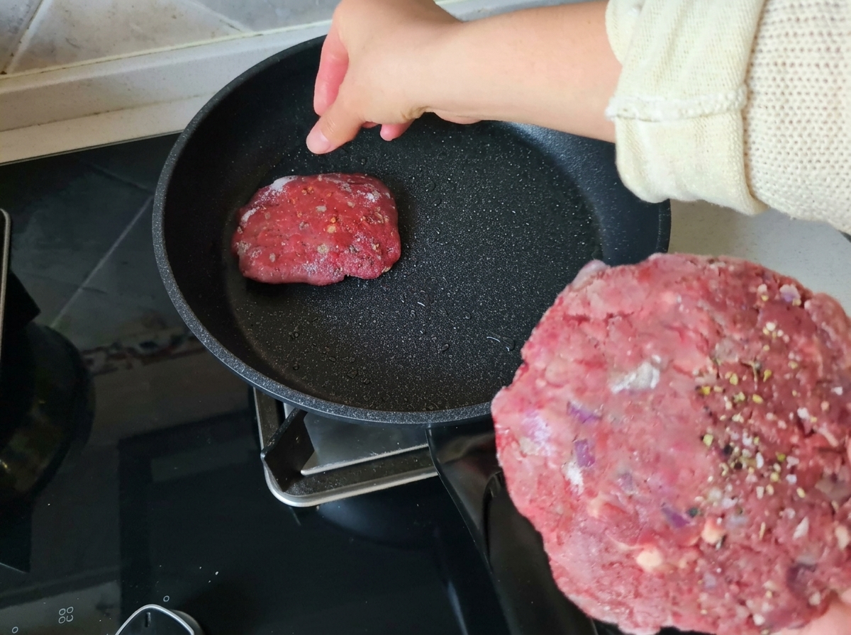 Une main tenant un steak de bœuf cru assaisonné tout en plaçant un autre steak dans une poêle antiadhésive chaude et légèrement huilée.