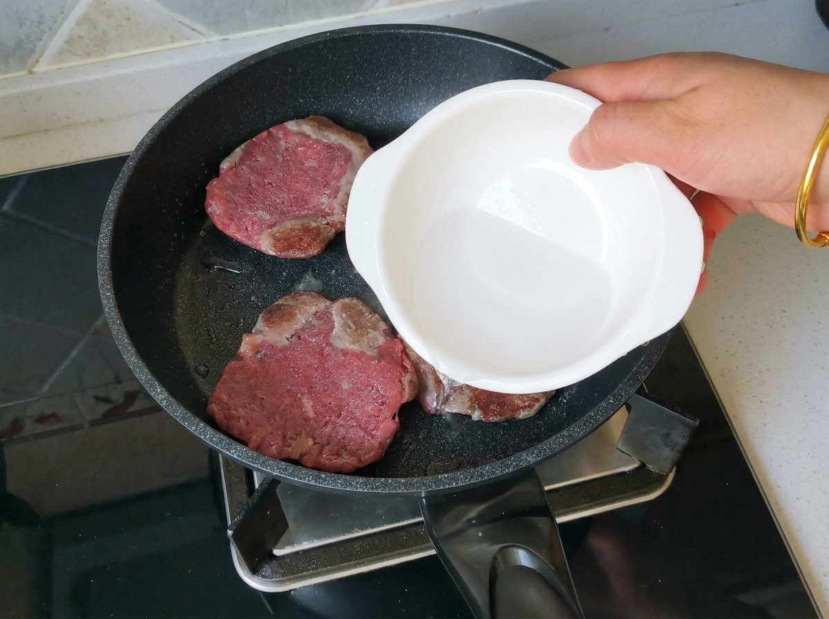 Une main versant un peu deau depuis un petit bol blanc dans une poêle contenant trois steaks de bœuf bien dorés.