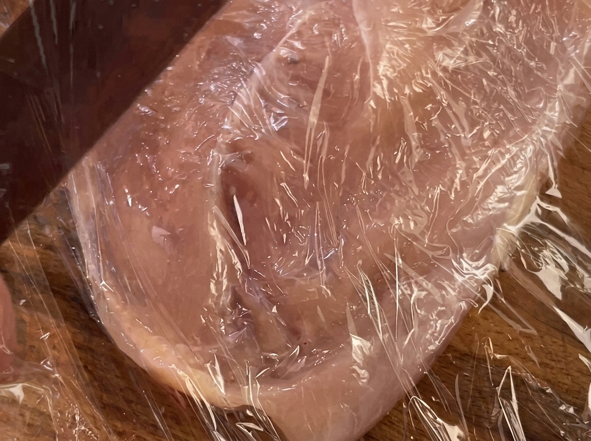 Un rouleau à pâtisserie pressant un blanc de poulet recouvert de film plastique.