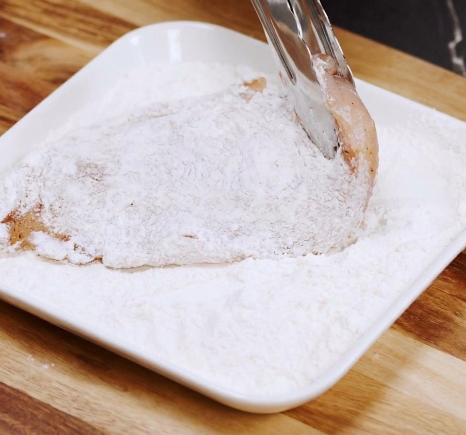 Filet de poulet cru enrobé de farine dans une assiette blanche avec des pinces en métal.