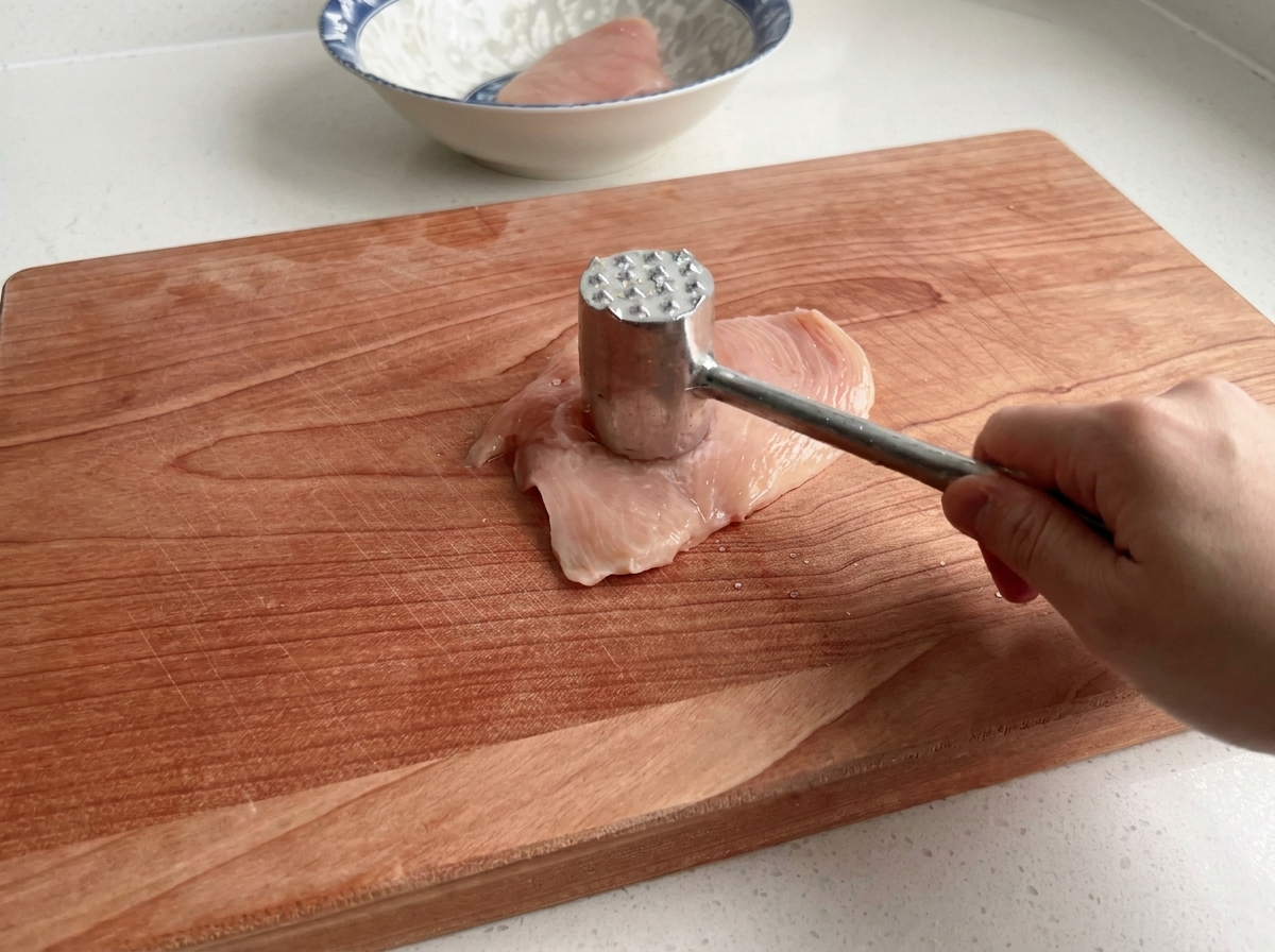 Gros plan dun maillet à viande attendrissant un blanc de poulet cru sur une planche en bois.
