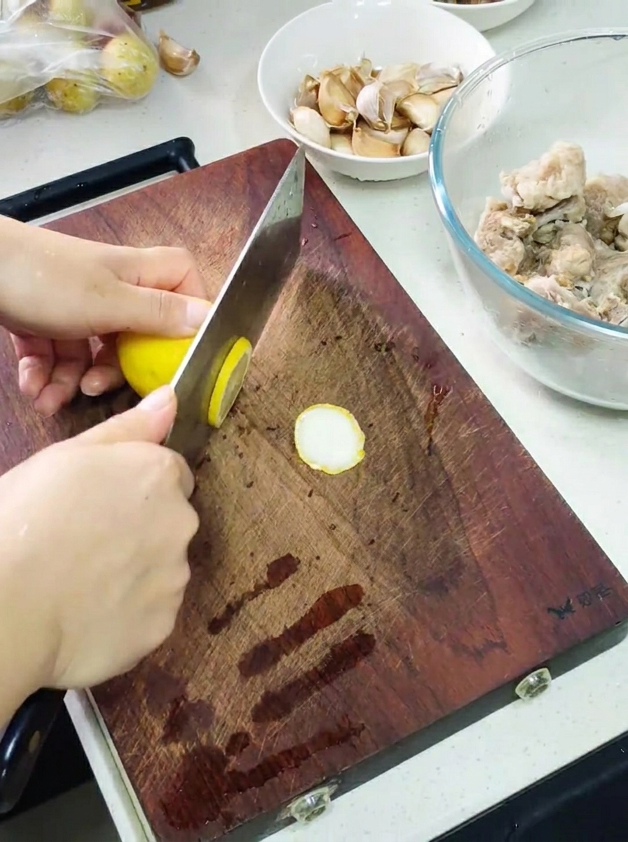 Mains utilisant un couperet pour trancher un citron sur une planche, avec un bol de travers marinés et des gousses dail à côté.