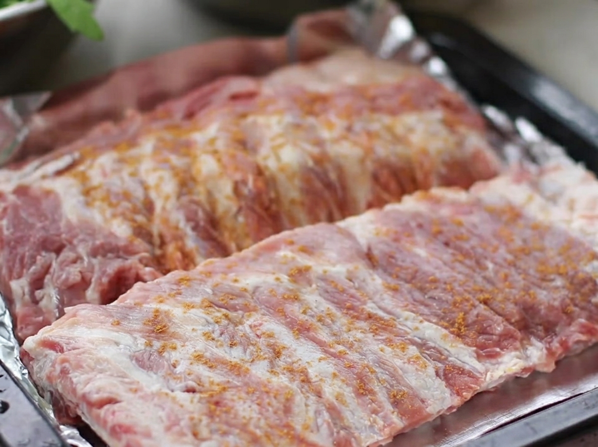 Travers de porc crus disposés sur une plaque de cuisson, généreusement recouverts dun mélange dépices, de poivre, de gingembre et dail.