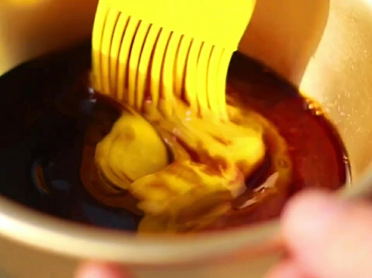 Un pinceau en silicone appliquant une sauce sombre et riche sur la surface de travers de porc partiellement cuits dans un plat.