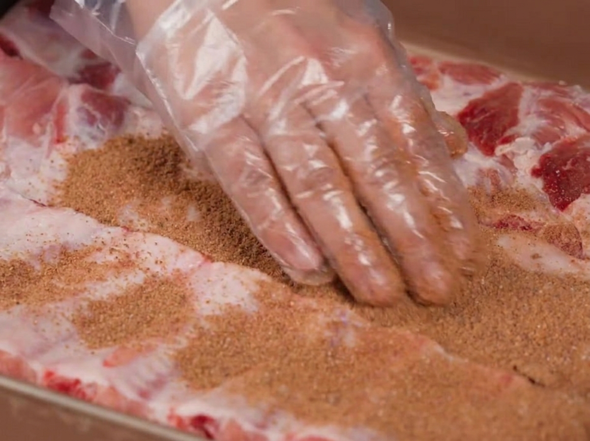 Mains portant des gants en plastique frottant un mélange dépices brun sur des travers de porc crus.