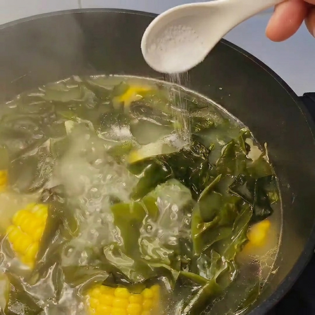 Une cuillerée de sel ajoutée à une casserole de soupe mijotante contenant du kombu et du maïs.