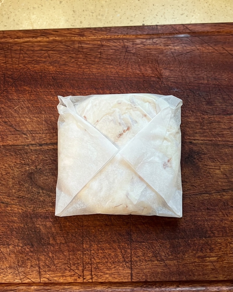 Une crêpe à loignon pliée et enveloppée dans du papier cuisson, en train dêtre pressée à plat sur une planche à découper en bois.