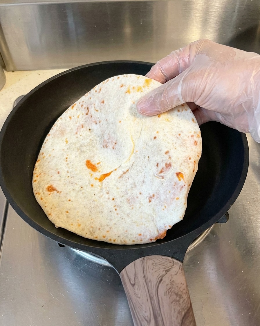 Une main gantée place une crêpe enveloppée dans du papier cuisson dans une poêle antiadhésive noire chaude.