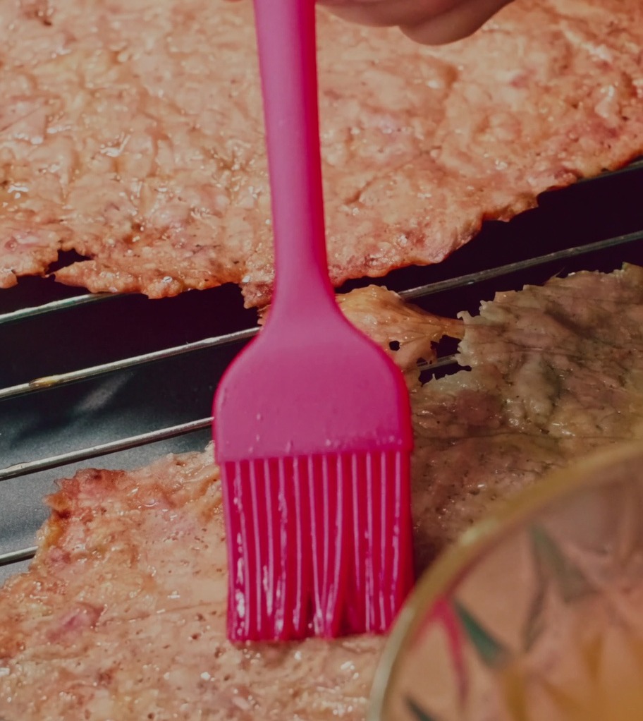 Un pinceau en silicone applique un glaçage miel-eau sur des tranches de jerky de porc sur une grille.