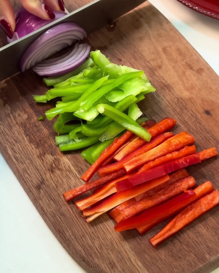 Tranches de poivron vert, poivron rouge et oignon rouge disposées proprement sur une planche en bois.